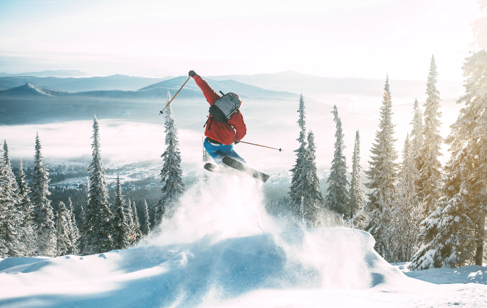 Imagem de jovem de costa fazendo um salto esquiando. Ao horizonte, temos montanhas e árvores cobertas de neve.