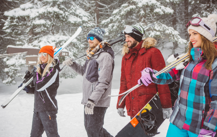 Imagem de quatro jovens, duas mulheres e dois homens, caminhando em uma paisagem coberta de neve. Cada um carrega seu próprio equipamento de esqui.