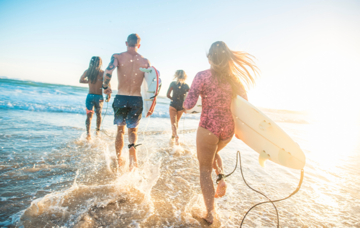 Imagem de quatro jovens, duas mulheres e dois homens, correndo em direção ao mar carregando pranchas de surfe em uma praia ensolarada.