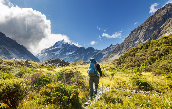 Imagem das montanhas da Nova Zelândia sob um céu azul com um jovem no centro, virado para as montanhas, fazendo trilha.