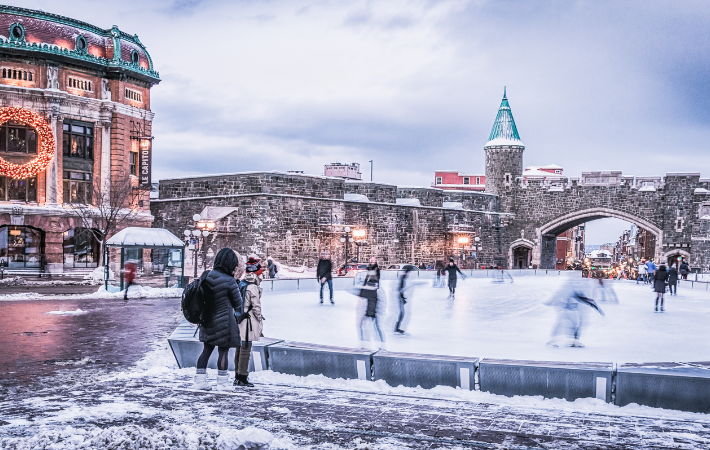 Foto de um Festival de Inverno europeu, a imagem apresenta um lago congelado com pessoas patinando, cercado por construções antigas cobertas de nev